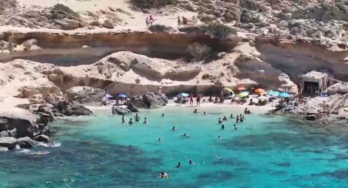 Bañistas caminando por el camino hacia es Caló des Mort mientras otros se protegen del sol bajo los voladizos