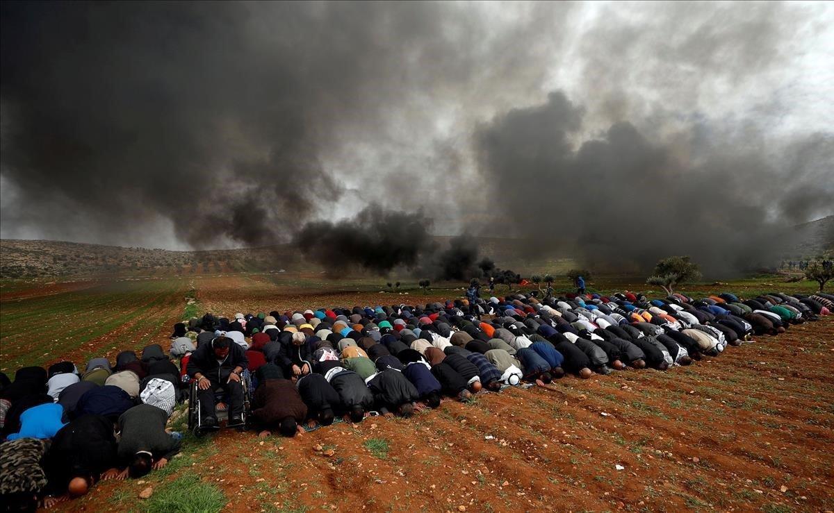 Los palestinos realizan las oraciones del viernes, mientras el humo se levanta al quemarse los neumáticos durante una protesta contra los asentamientos judíos en la aldea de al-Mughayer cerca de Ramallah en la Cisjordania ocupada por Israel.