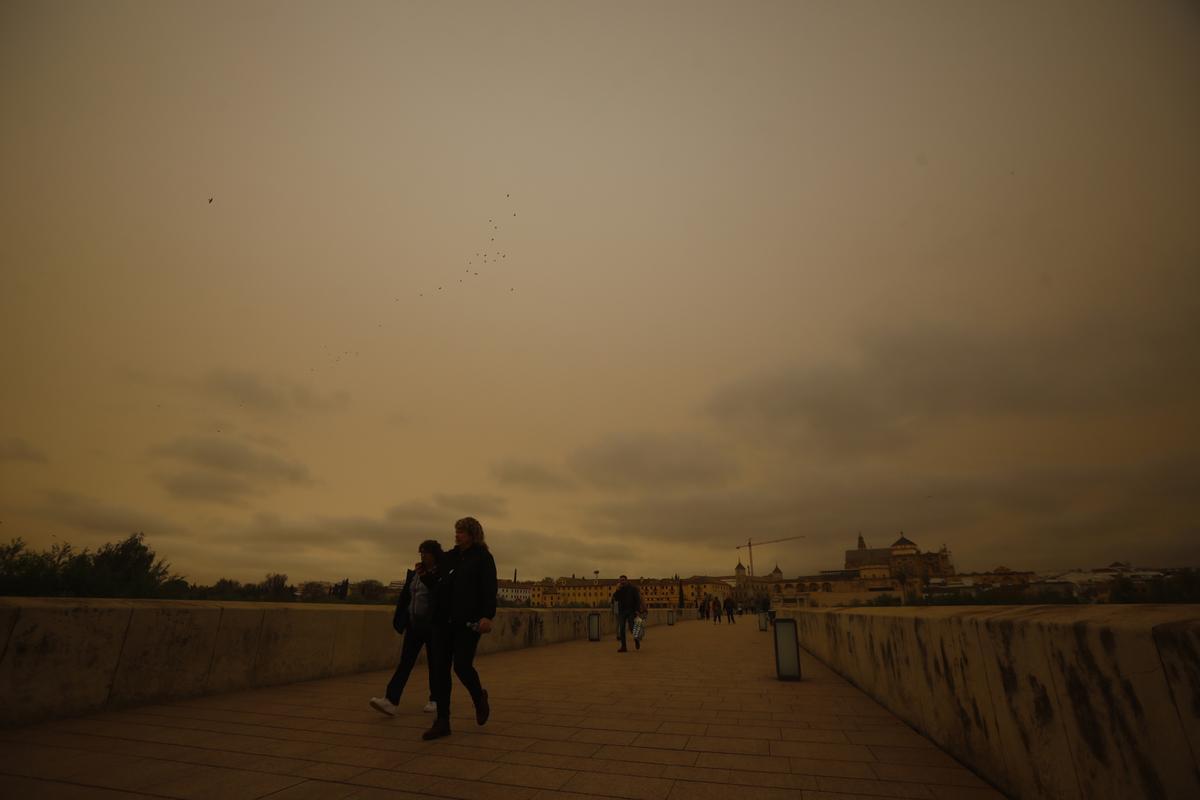 Varias personas caminan por el Puente Romano de Córdoba, bajo un cielo cubierto por la calima.
