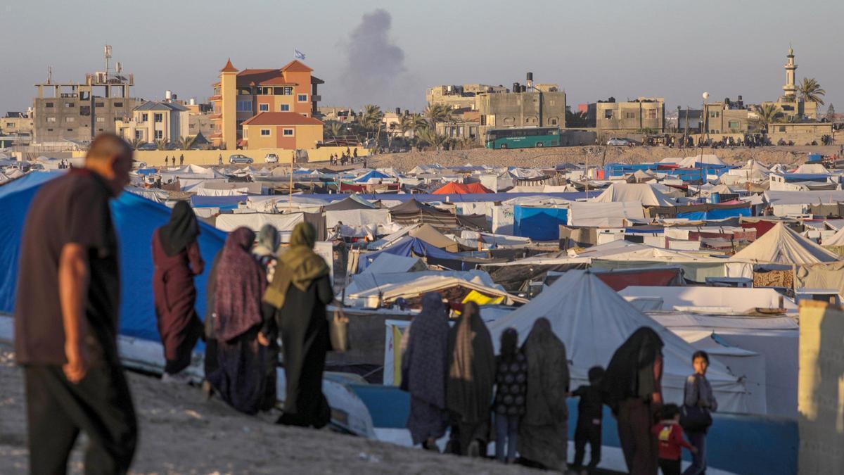 Palestinos desplazados, en un campo de acogida mientras a lo lejos el humo de un ataque israelí se eleva sobre Rafah