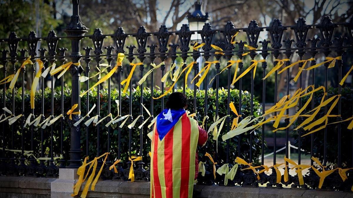 Un joven con la bandera indepentista anudada a su espalda coloca lazos amarillos en la verja que rodea el Parc de la Ciutadella, el 17 de enero del 2018