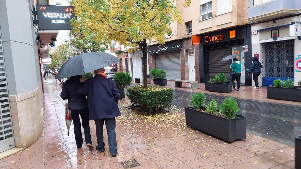 Varias personas pasean, bajo los paraguas, por una céntrica calle de Alzira.