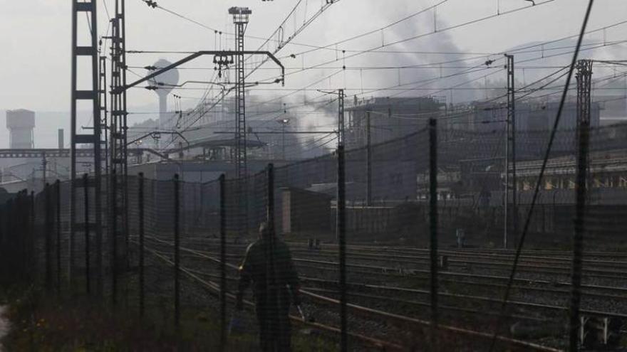 Un hombre camina junto a las vías del tren cerca del puerto, con un paisaje de fábricas humeantes al fondo.