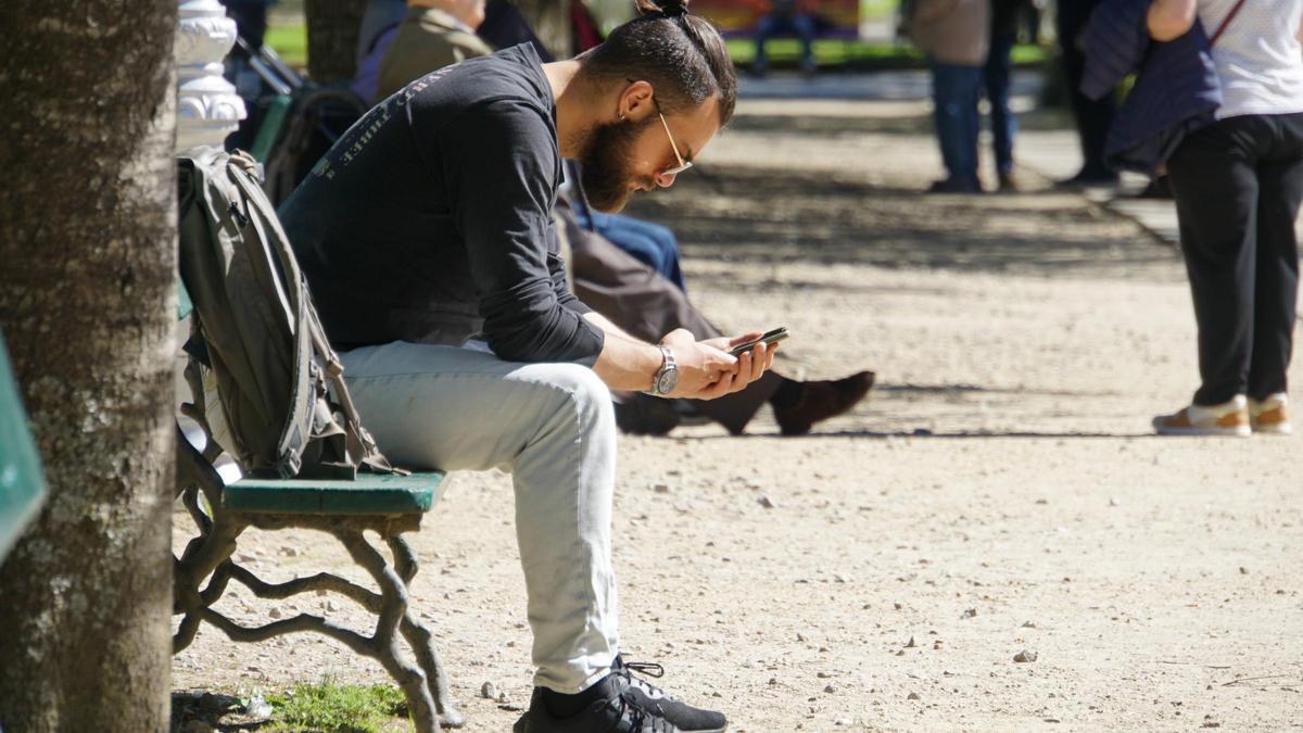 Un joven consulta su teléfono móvil sentado en un banco del parque de la Alameda
