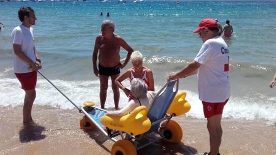 Personal de la Creu Roja portant una dona amb una cadira amfíbia en una platja gironina.