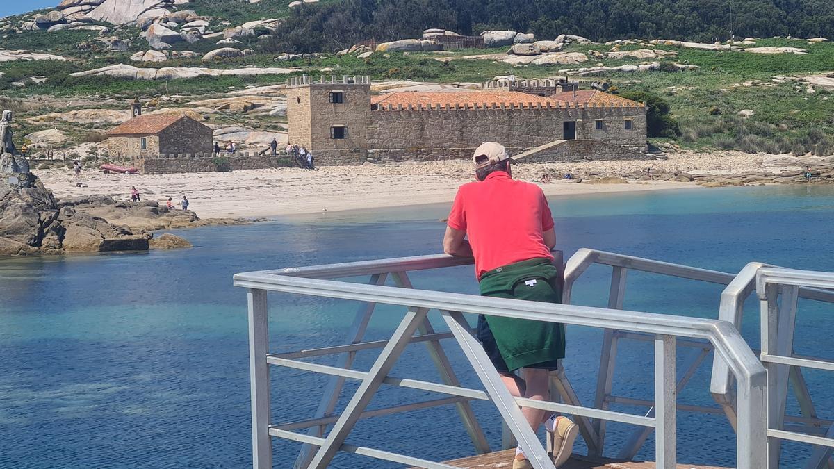 Un hombre observa desde el embarcadero una de las playas y las edificaciones de la capilla y el viejo pazo.