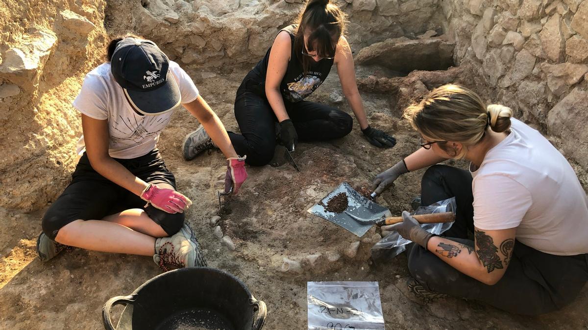 En primer terme, unes arqueòlogues excavant una llar de foc; al fons, l&#039;estructura de la cuina grega del segle VI aC. Foto publicada el 7 de desembre del 2021 (Horitzontal)