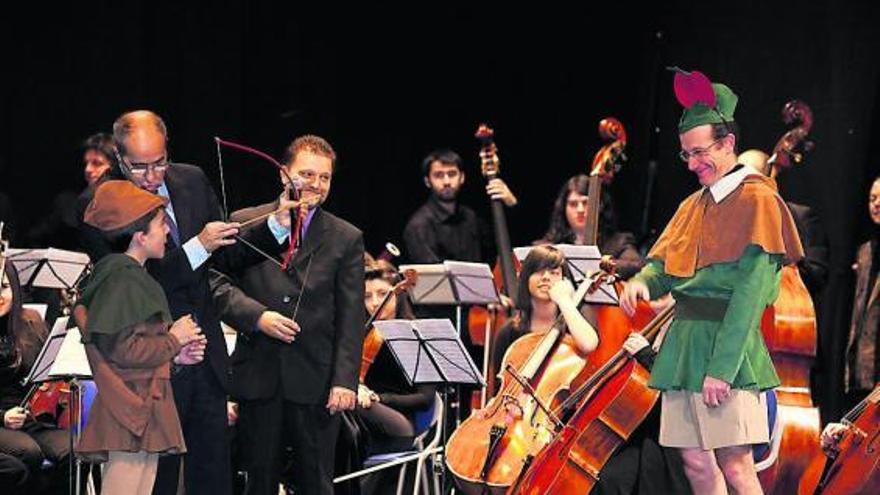 Fernando Argenta y Manuel Paz enseñan a un niño a disparar con arco, con la Orquesta de Siero al fondo.