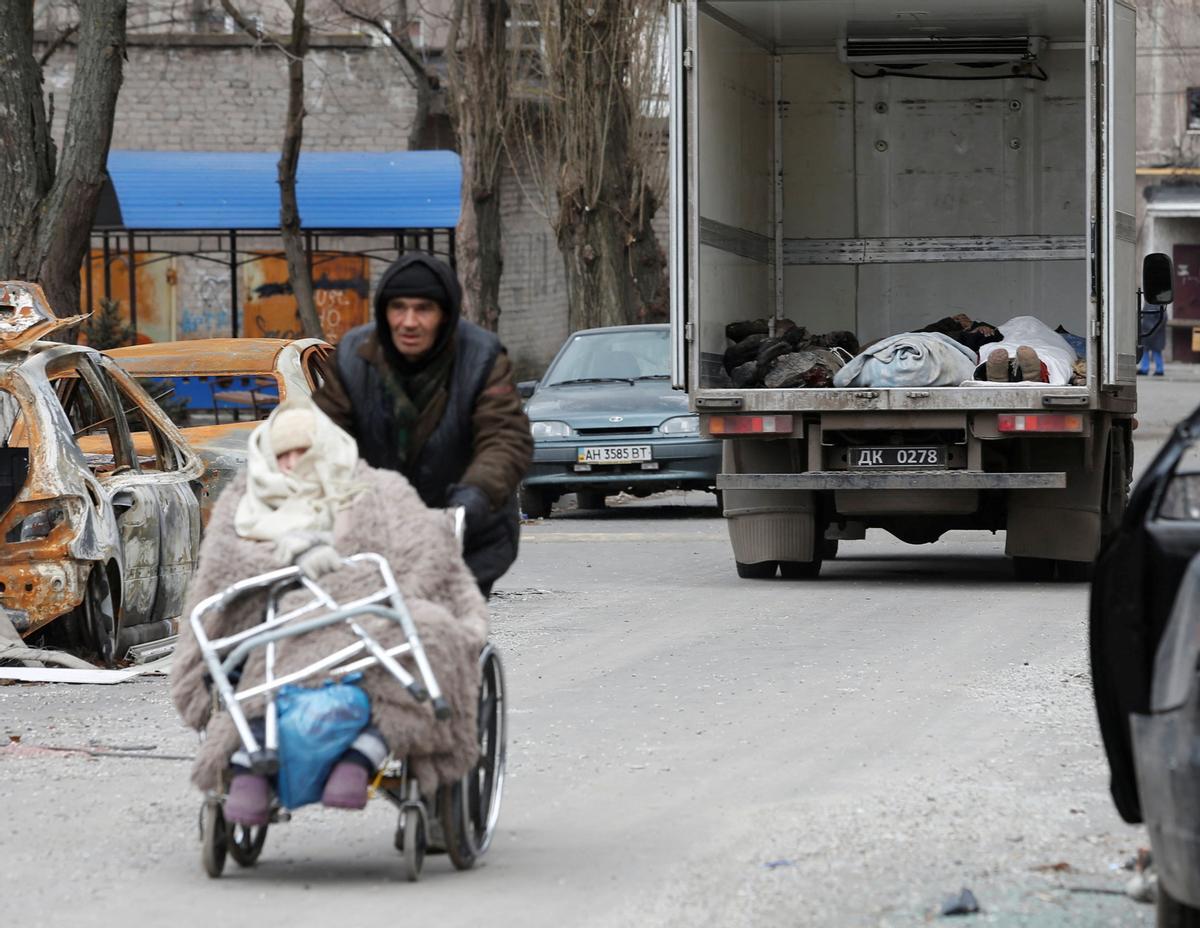 Un hombre empuja a una anciana en silla de ruedas, mientras los cuerpos de los muertos recogidos por miembros del servicio de las tropas prorrusas se ven dentro de un vehículo antes de ser transportados a la morgue durante el conflicto Ucrania-Rusia en el puerto del sur ciudad de Mariupol.