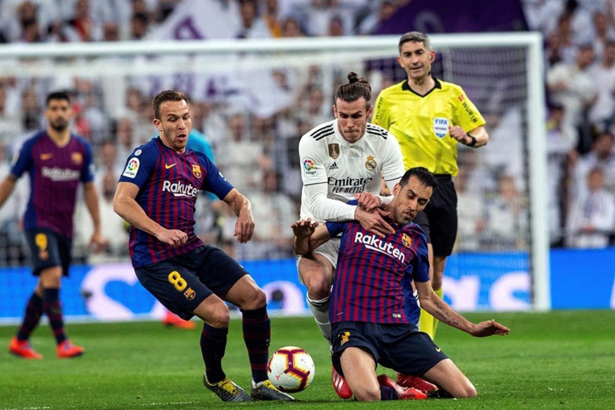 Gareth Bale (c), lucha por el balón ante los jugadores del FC Barcelona, Arthur Melo (i) y Sergio Busquets durante el partido entre el Real Madrid y el FC Barcelona en el Estadio Santiago Bernabéu de la jornada 26 de La Liga