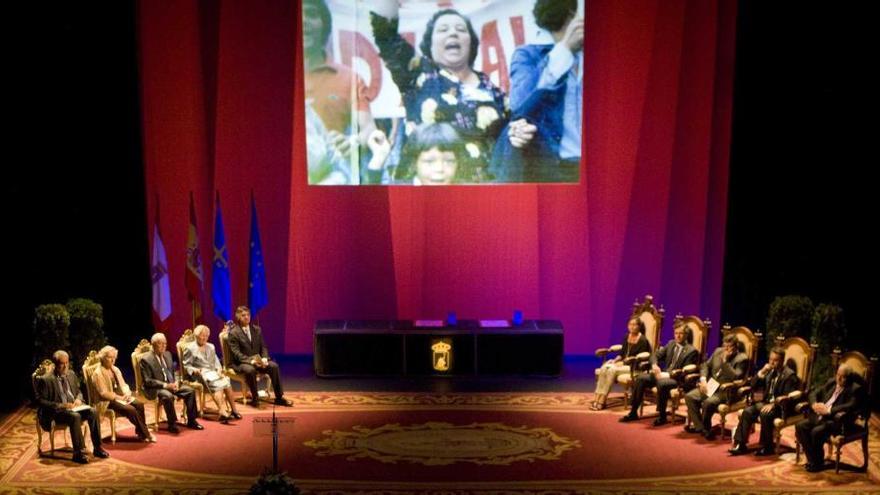 El escenario del teatro Jovellanos, con los galardonados, a la izquierda, y la Alcaldesa y los cuatro portavoces de los grupos políticos, a la derecha; en la imagen proyectada, María González.