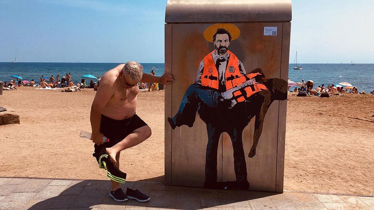 Un bañista se limpia la arena de los pies junto al dibujo de Òscar Camps pintado por Tvboy en la Barceloneta