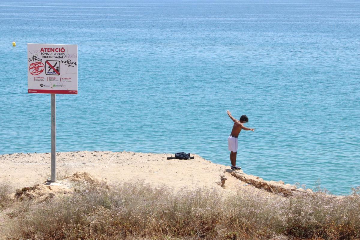Un menor procede a saltar desde las rocas de la playa del Miracle en Tarragona, ante un cartel de prohibición.