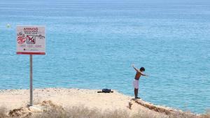 Un menor procede a saltar desde las rocas de la playa del Miracle en Tarragona, ante un cartel de prohibición.