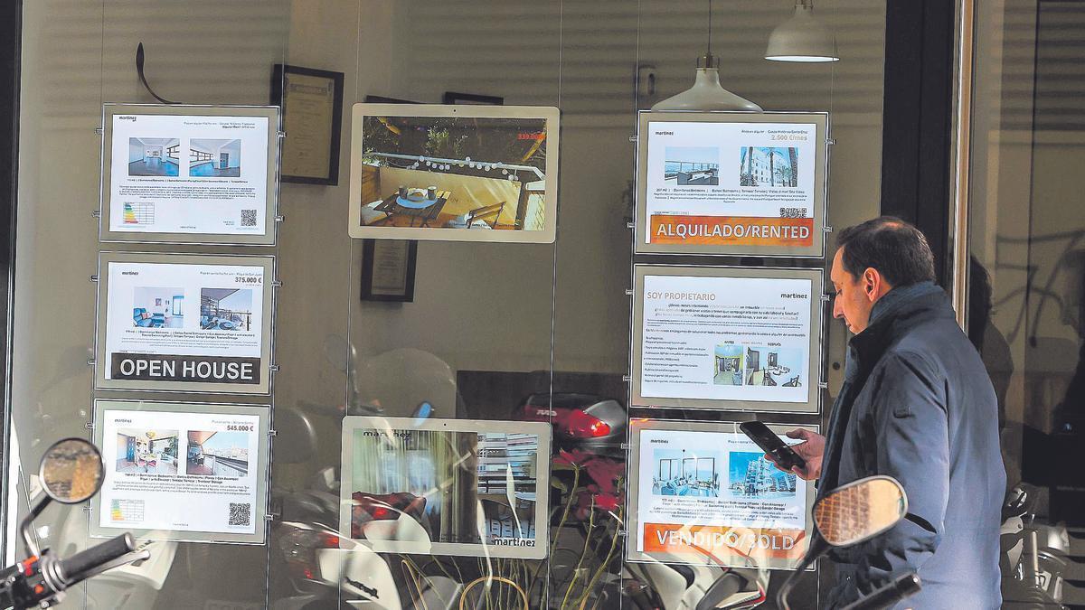 Un hombre consulta su móvil frente a las ofertas de alquiler y venta de pisos de una inmobiliaria de Alicante.