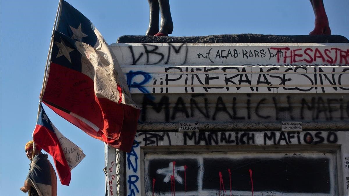 Un manifestante con una bandera de Chile pasa junto a la base de una estatua ecuestre repleta de pintadas.