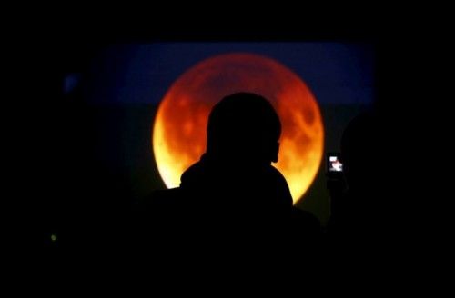 People look at the screen displaying the moon, appearing in a dim red colour, which is covered by the Earth's shadow during a total lunar eclipse in Warsaw