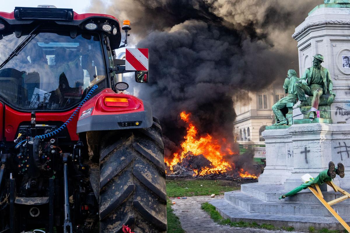 Varios manifestantes queman basura junto a una estatua y un tractor frente al Parlamento Europeo. La protesta de agricultores denuncia el acuerdo con Mercosur y la reforma de los subsidios de la PAC por la amenaza que suponen para el sector comunitario. 18 de diciembre de 2025, Bruselas.