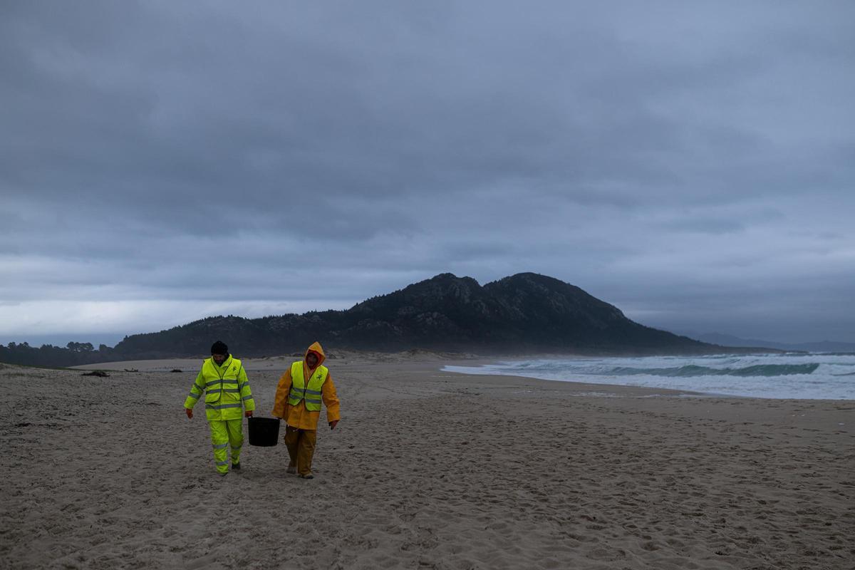Dos hombres cargan un cubo lleno de pellets, en la playa Area Maior, a 13 de enero de 2024, en Muros, A Coruña, Galicia (España).