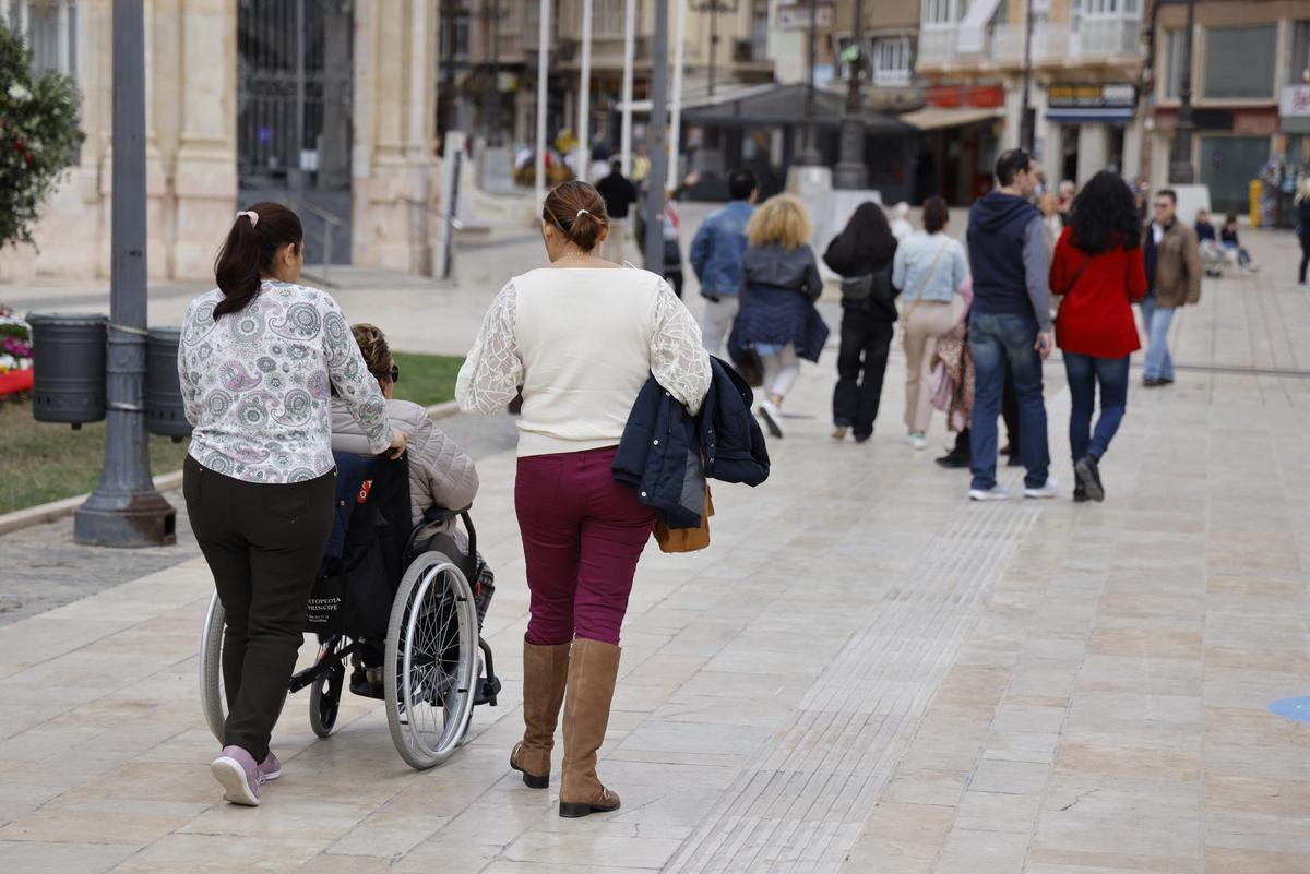 Una cuidadora, acompañada de una amiga, empuja la silla de ruedas de una mujer dependiente en Cartagena.