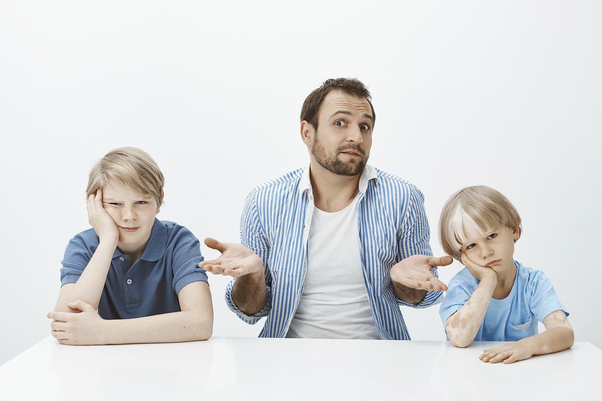 Portrait of confused unaware european father sitting with sons at table, shrugging with spread palms and questioned expression while sons being bored or offended over grey background, Portrait of confused unaware european father sitting with sons at table, shrugging with spread palms and questioned expression while sons being bored or offended over grey background