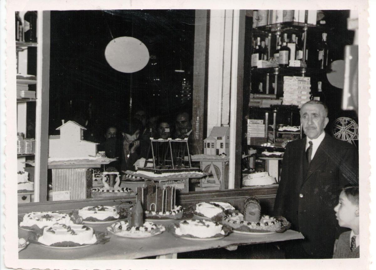 José de Blas, el artífice del carbayón, junto a uno de sus nietos ante varias tartas de Pascua, que observan al otro lado del ventanal, varias personas.