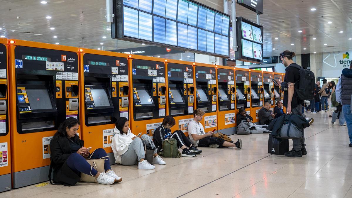 Máquinas expendedoras de tickets fuera de servicio y pasajeros de los trenes de Rodalies de Renfe matando el tiempo como pueden en la estación de Sants por el parón de la actividad a consecuencia de la alerta de protección civil con motivo de las fuertes lluvias en el territorio