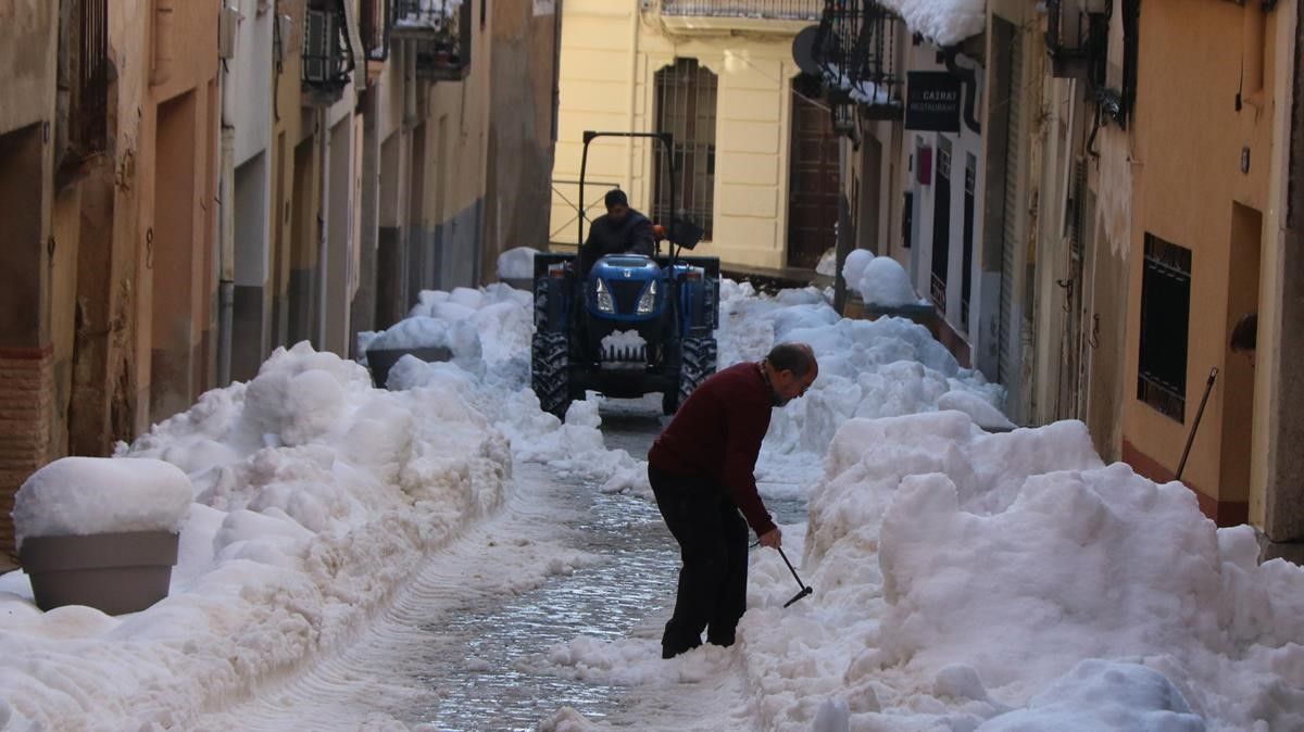 Pla obert d un carrer de Falset nevat amb un vei retirant neu de la porta de casa seva i un tractor netejant  Imatge de l 11 de gener de 2021  (Horitzontal) Roger Segura ACN