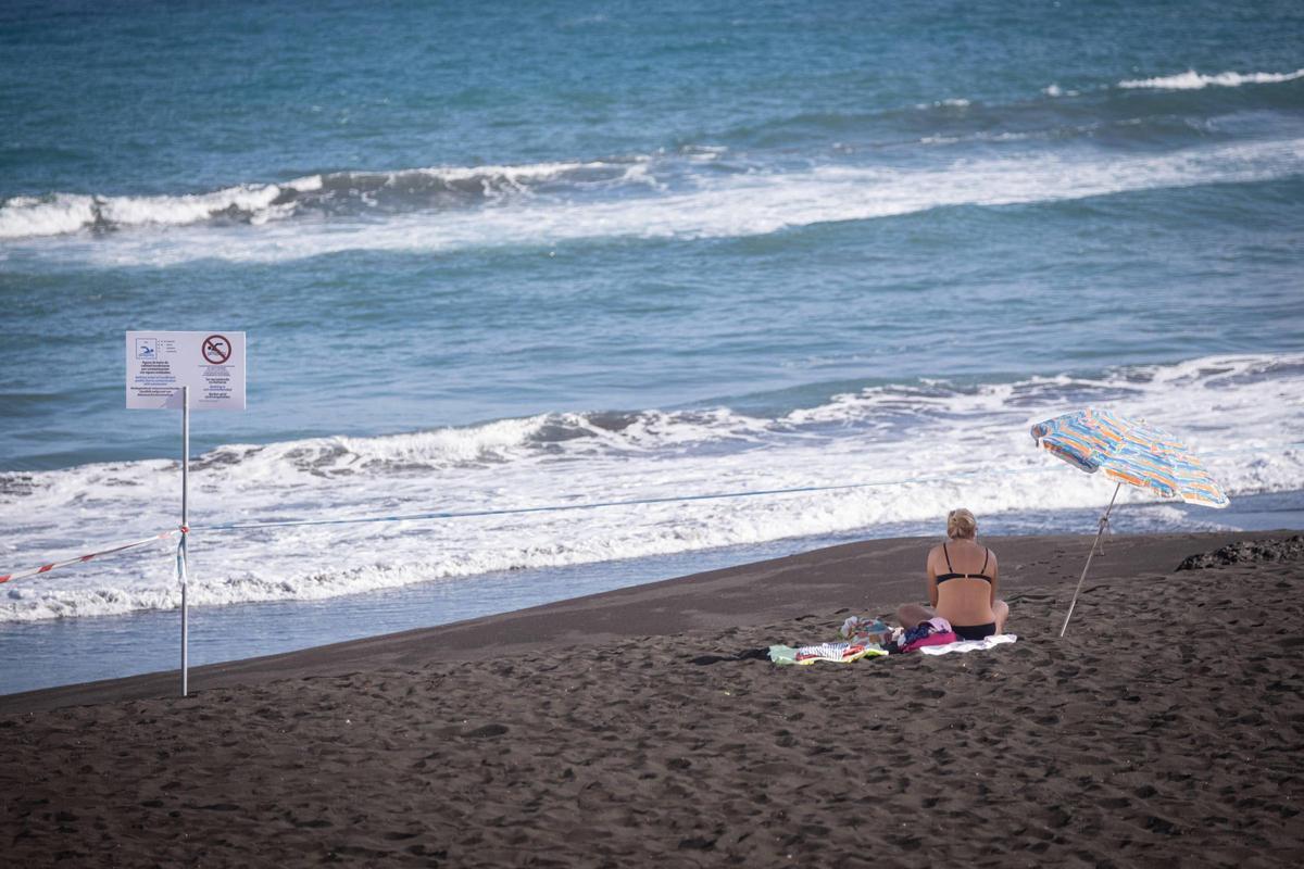 Una mujer toma sol en Playa Jardín y al lado carteles que informan de la prohibición de bañarse en sus aguas.