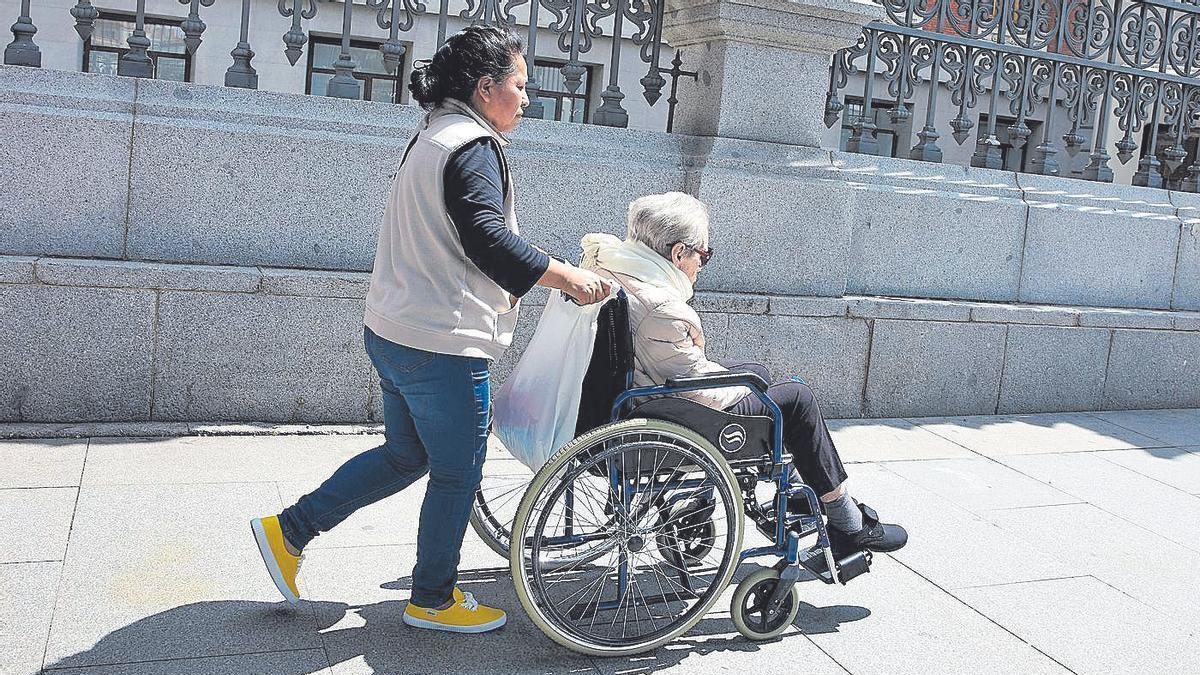Una mujer mayor en silla de ruedas y su cuidadora pasean por una calle, en una fotografía de archivo.