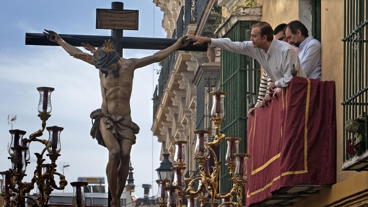 Un hombre asomado a un balcón alcanza a tocar la mano del Cristo de La Salud de la Hermandad de San Bernardo.