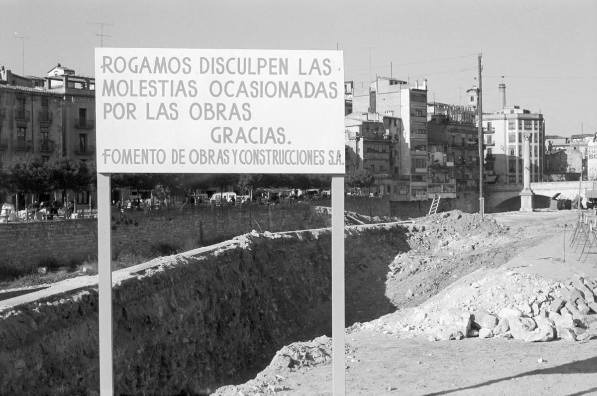 Cartell anunciant les obres de construcció d'una plataforma sobre el riu Onyar sobre la qual s'ubicarà l'actual plaça Catalunya. Al fons a la dreta s'observen el Pont de Pedra, la torre de la Casa de les Aigües i la xemeneia de la Fàbrica Grober