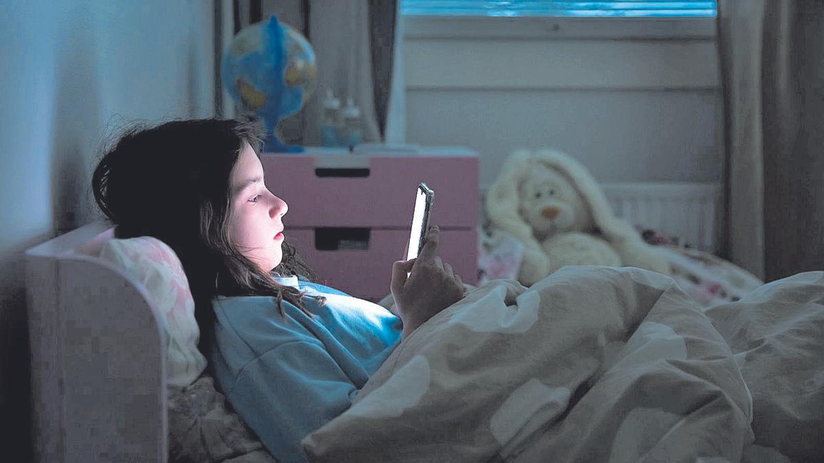 Una niña mira la pantalla de un teléfono móvil, tumbada en una cama.