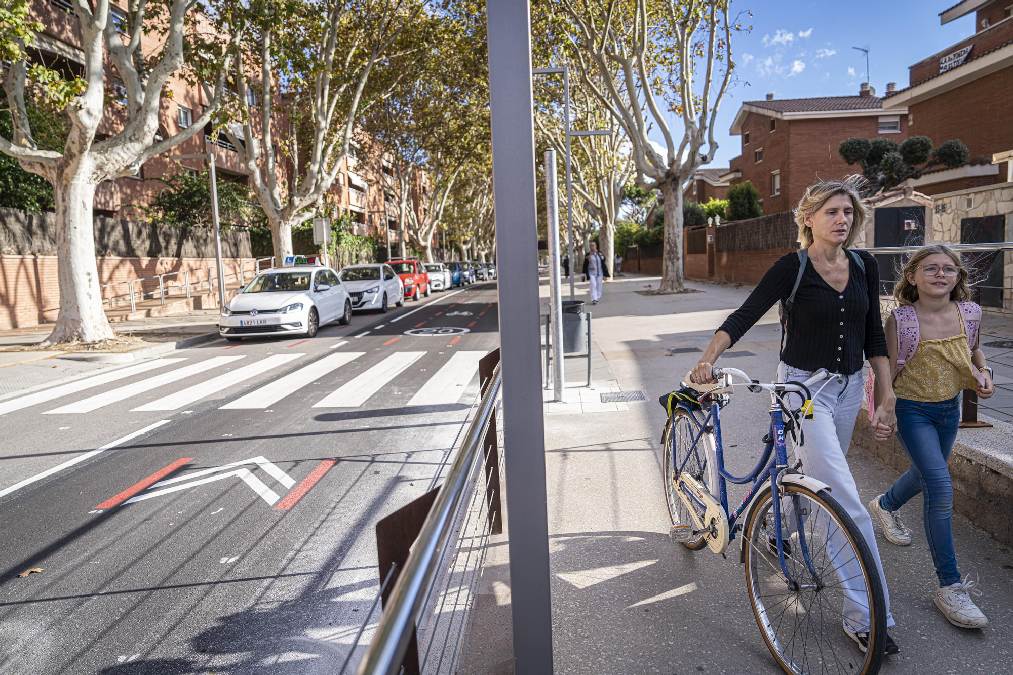Una madre y su hija caminan con su bici por la acera peatonal de la Avenida de la Pineda de Castelldefels