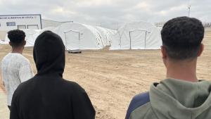 Tres jóvenes migrantes esperan en la mañana de este lunes para ser alojados en el nuevo campamento temporal que se está habilitando en el muelle de Poniente del puerto de Ceuta que previsiblemente entrará en funcionamiento mañana martes. 