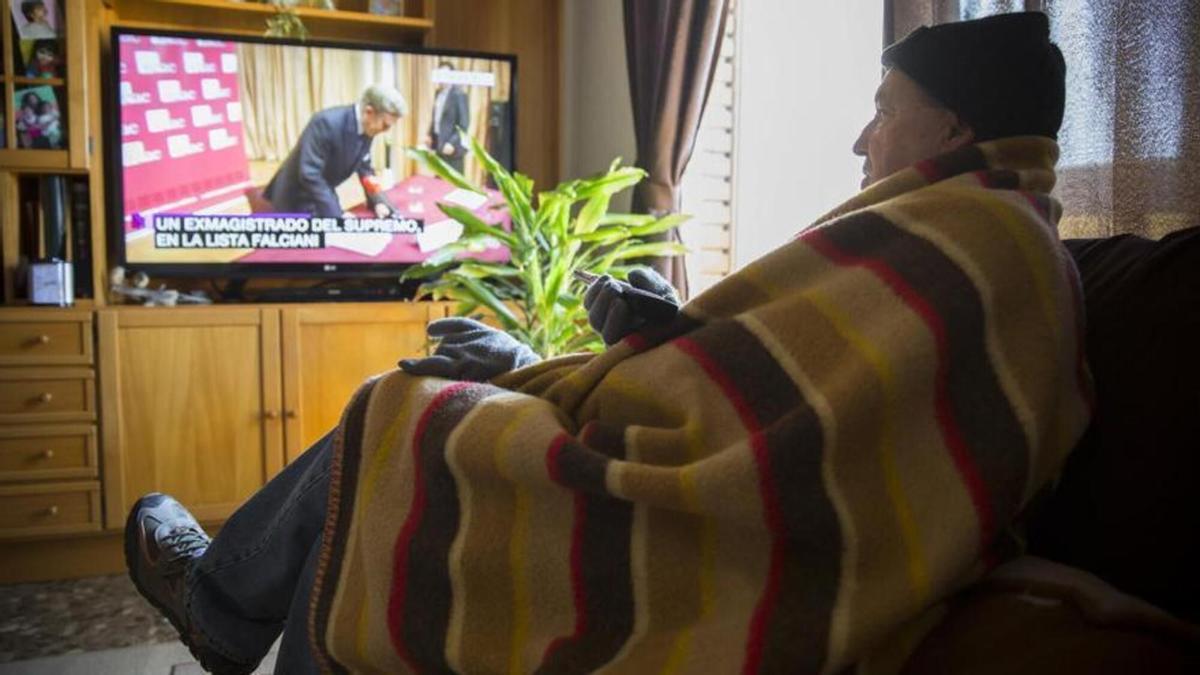 Un hombre ve la televisión tapado con una manta y guantes para reducir el consumo eléctrico del hogar.