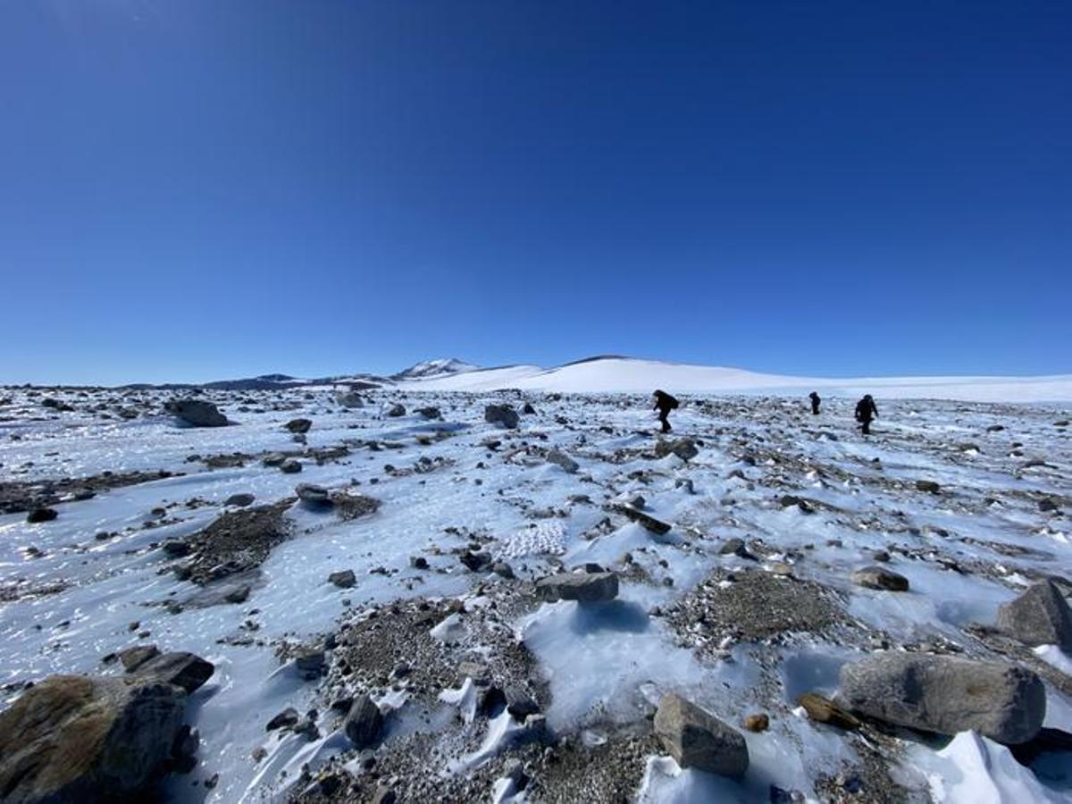 Rocas esparcidas por un campo de hielo, con los científicos buscando meteoritos en el fondo.