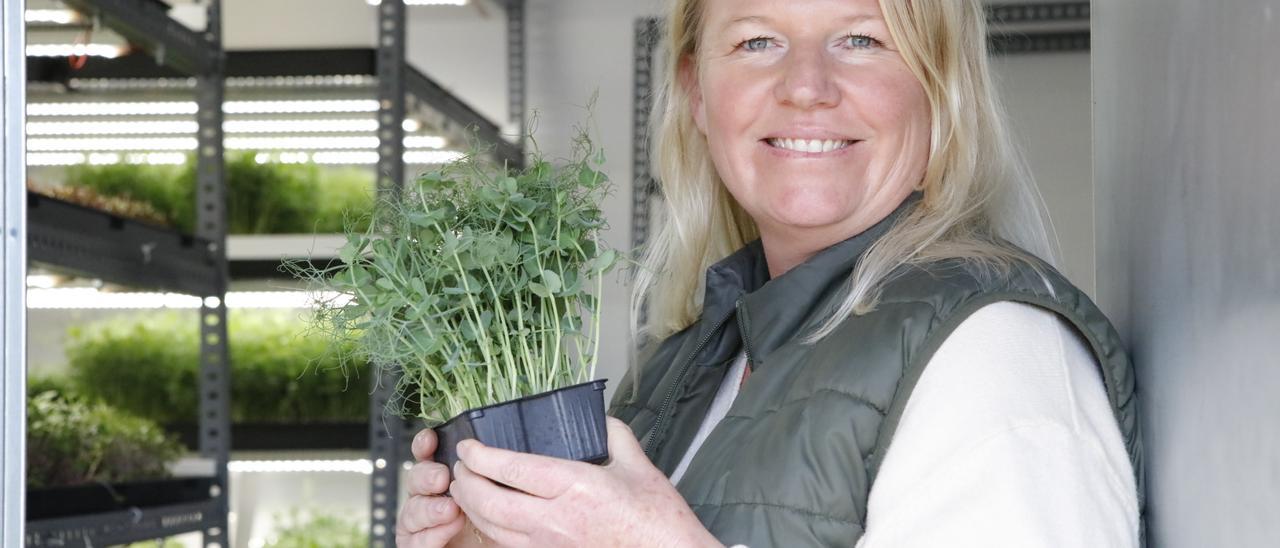 Becky Sell mit einer Pflanzschale, in der mit krausen Stängeln die Erbse wächst, im Hintergrund die ökologischen Kräuterzucht.