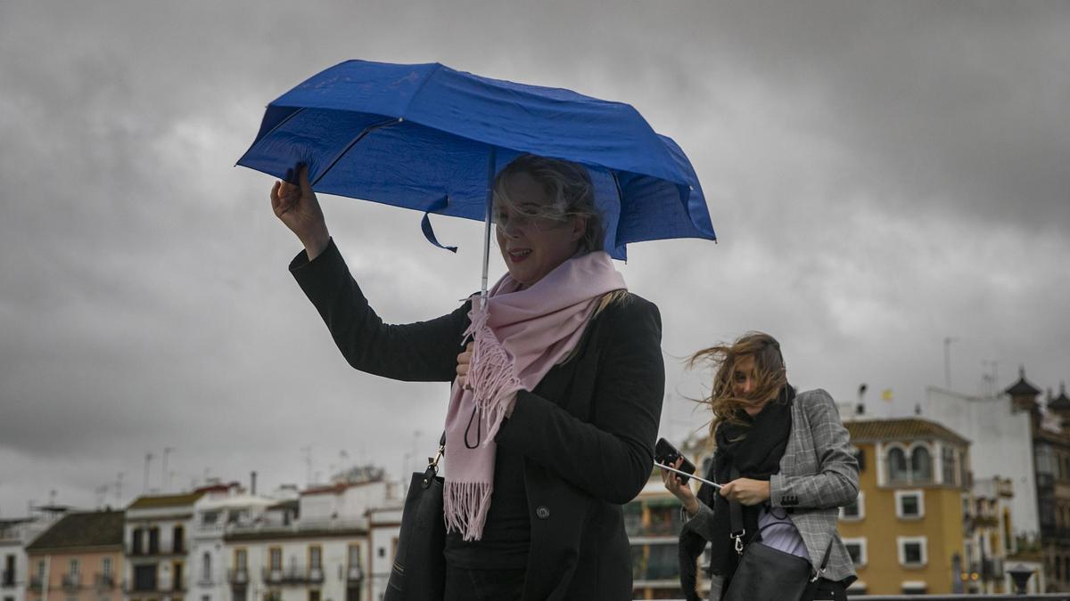 Archivo - Dos mujeres intentan protegerse del viento con su paragüas mientras caminan por el Puente de Isabel II, en Sevilla (Andalucía).
