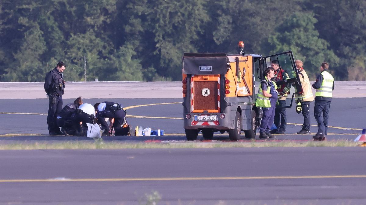 Sicherheitspersonal und Polizisten stehen auf dem Flugfeld und versuchen Aktivisten der Gruppe Letzte Generation vom Asphalt zu lösen, nachdem sie sich festgeklebt haben.