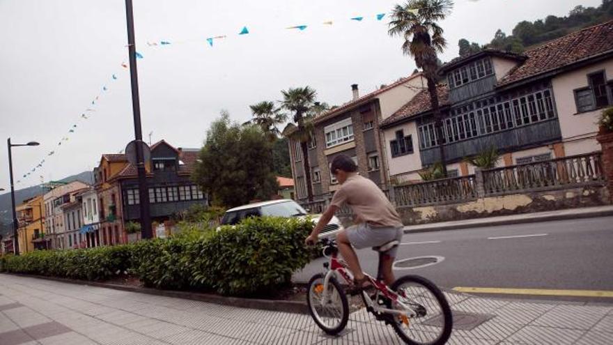 Un niño pasa en bicicleta por la avenida Príncipe de Asturias de Olloniego. A la derecha, una perspectiva de Tudela Veguín, con la fábrica de cemento al fondo.