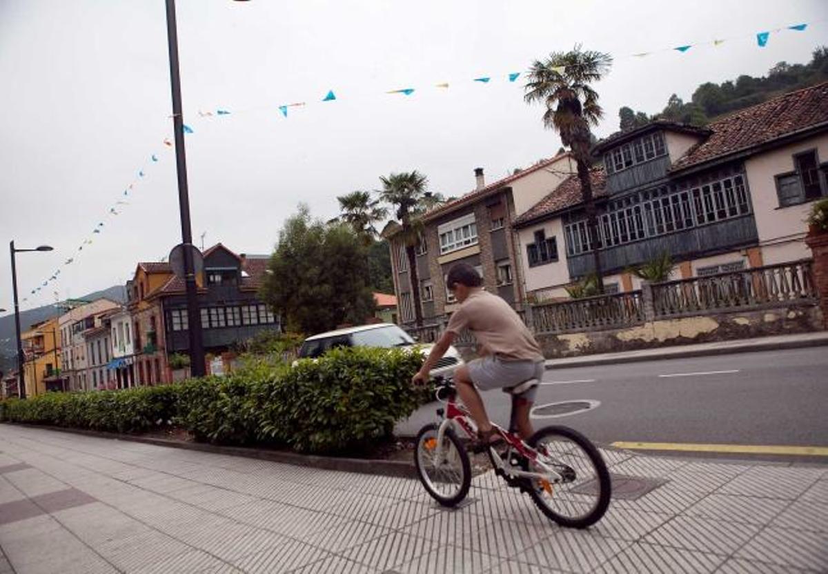 Un niño pasa en bicicleta por la avenida Príncipe de Asturias de Olloniego. A la derecha, una perspectiva de Tudela Veguín, con la fábrica de cemento al fondo.