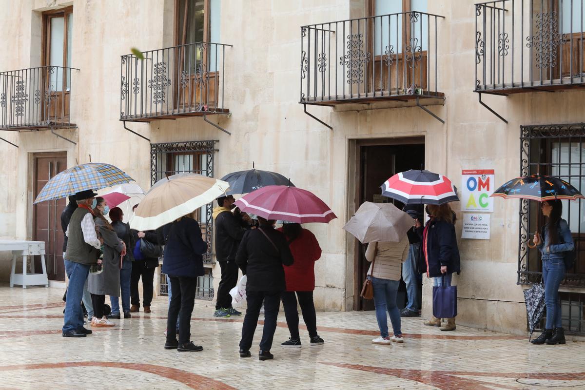 Gente esperando con paraguas a las puertas de la OMAC de la Plaça de Baix ante la falta de espacio dentro