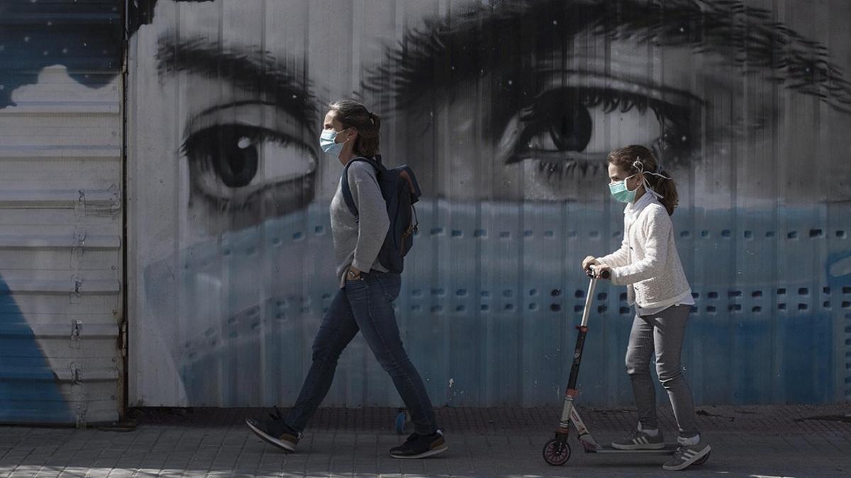 Una mujer y su hija pasean junto al grafiti de una joven con mascarilla, en Barcelona, el 28 de abril