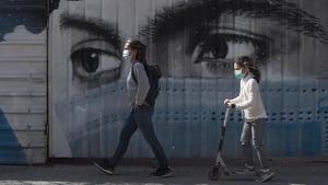 Una mujer y su hija pasean junto al grafiti de una joven con mascarilla, en Barcelona, el 28 de abril. 