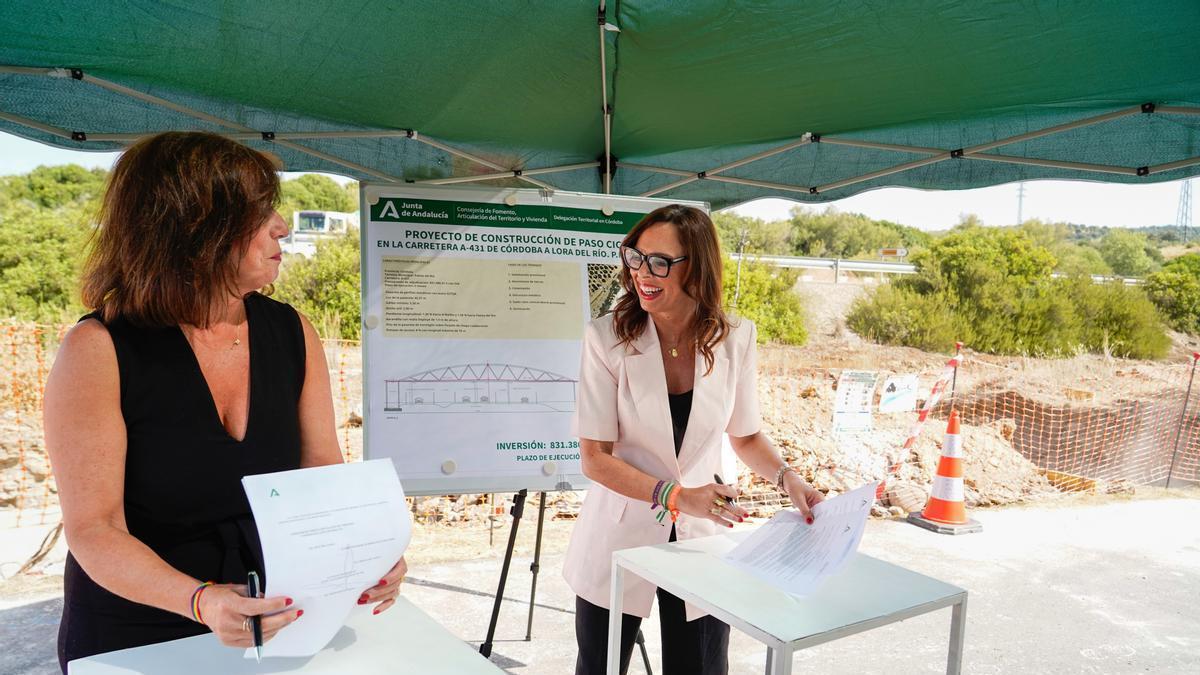 Rocío Díaz (dcha.) firma con Matilde Esteo, junto a las obras de la pasarela ciclopeatonal, el acuerdo de cesión de tramos de carreteras autonómicas al Ayuntamiento de Palma del Río en su paso por el municipio.