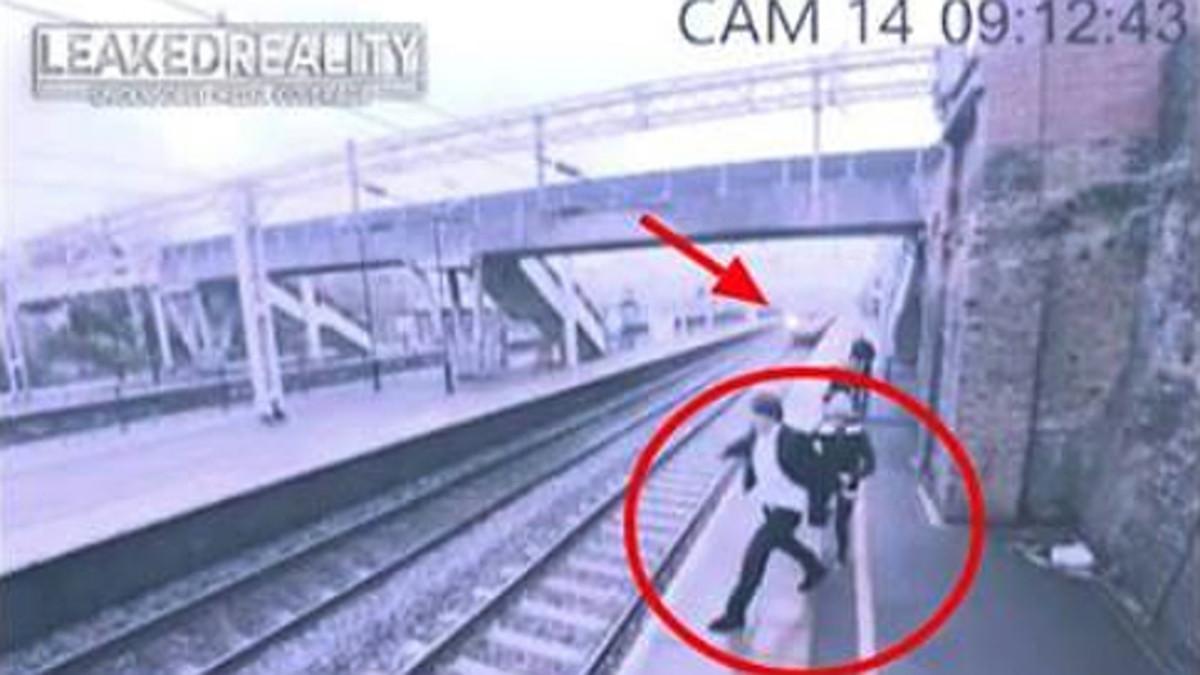 Momento en el que la mujer, detrás del hombre, le agarra del brazo y evita que se tire a la vía de tren.