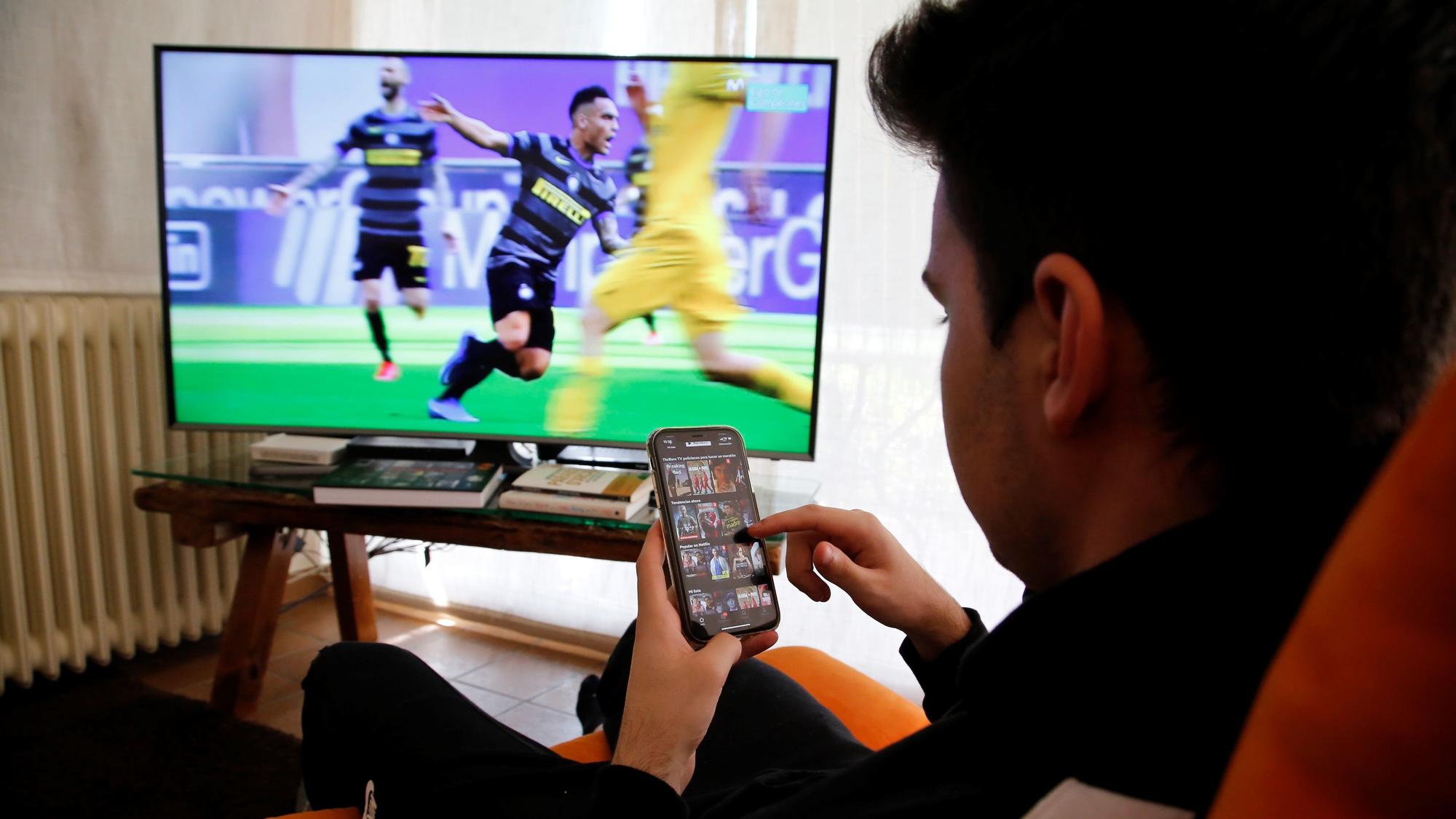 Un joven mira el móvil mientras en la televisión retransmiten un partido de fútbol.