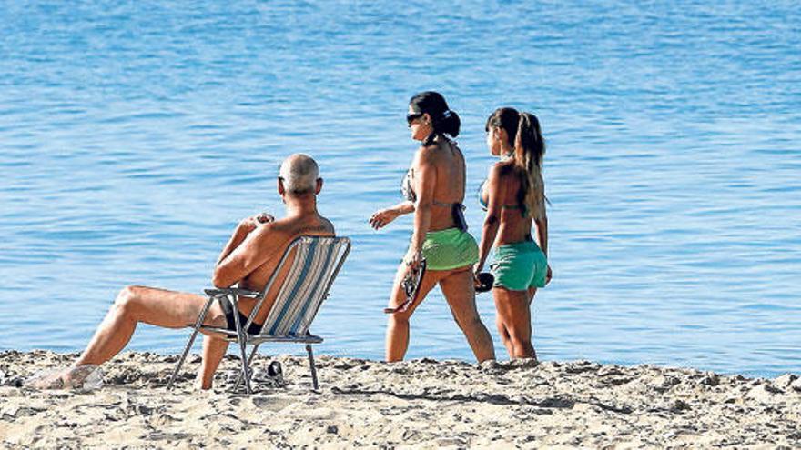 Dos chicas pasean por la orilla de la playa de Can Pere Antoni mientras un hombre se relaja antes de que llegue más gente.