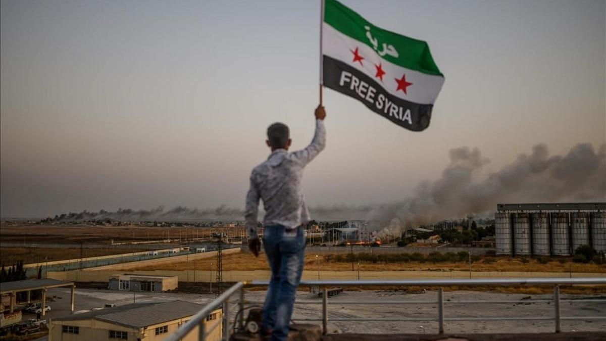 Un hombre sostiene una bandera en la que pide la liberación de Siria mientras las columnas de humo se elevan en las localidades fronterizas bombardeadas este domingo.